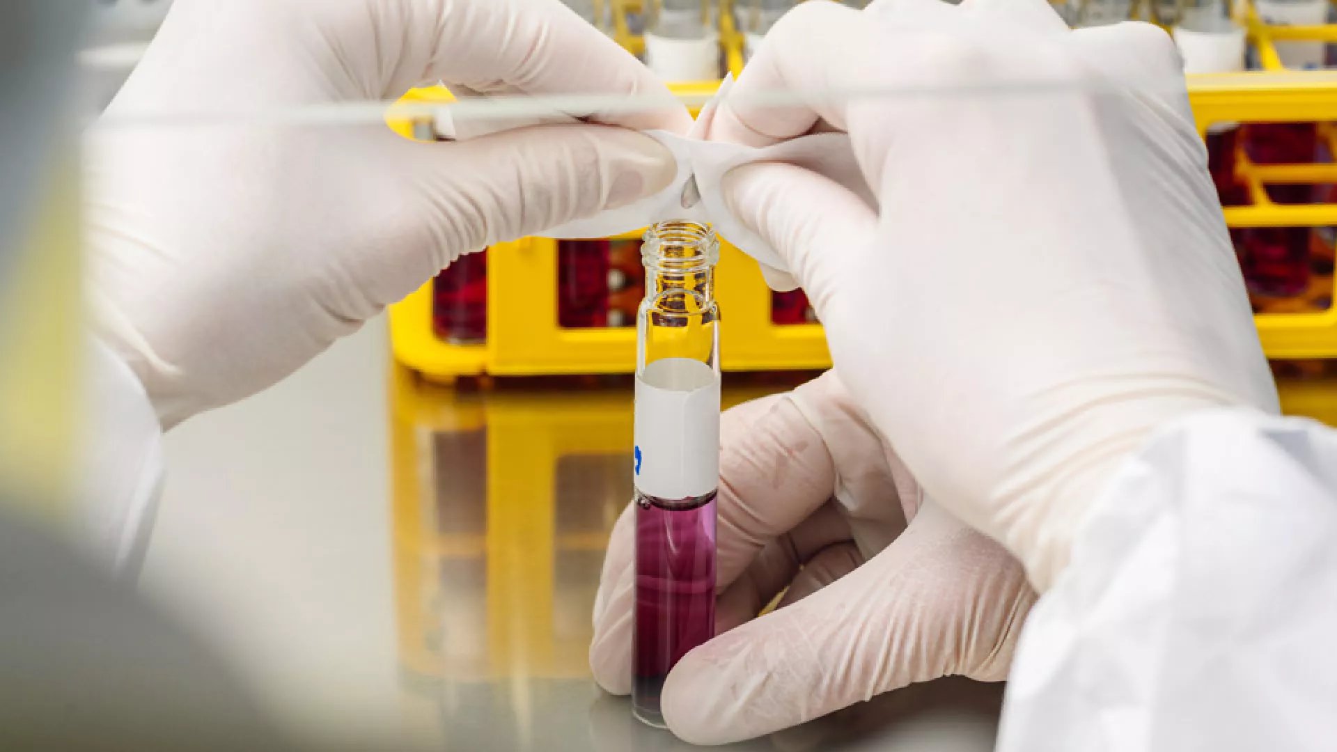 Gloved hands are shown handling a vial with a red liquid in a laboratory setting.